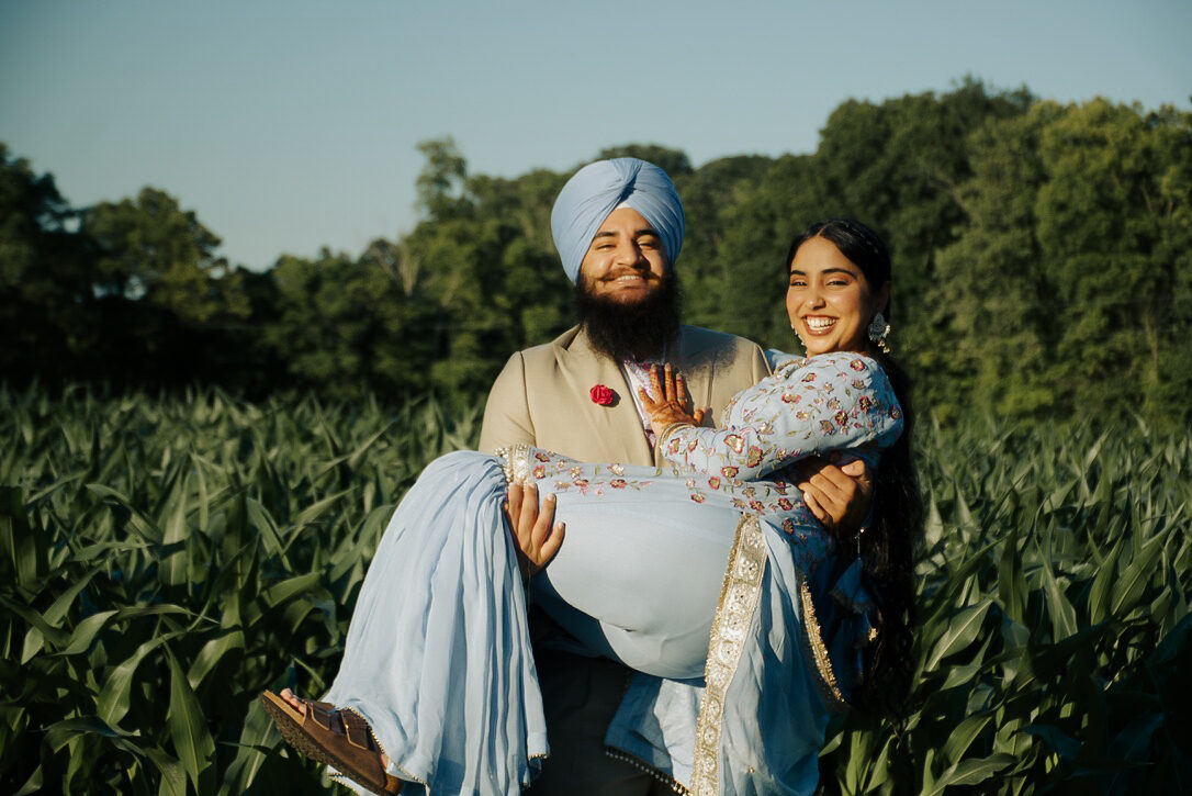 A groom carrying his partner through a cornfield at golden hour, both smiling.