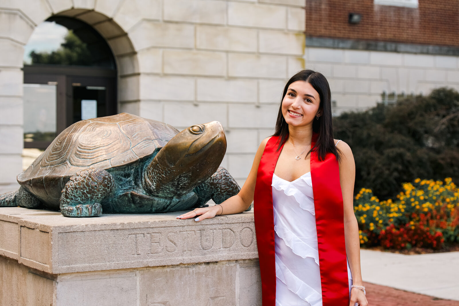 A graduate in a white dress and red stole seated by the Testudo statue.