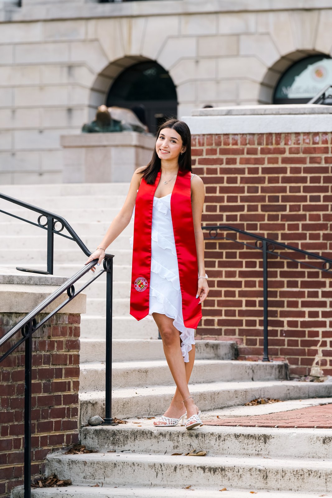 A graduate in a white dress and red stole standing on stone steps beside a brick archway.