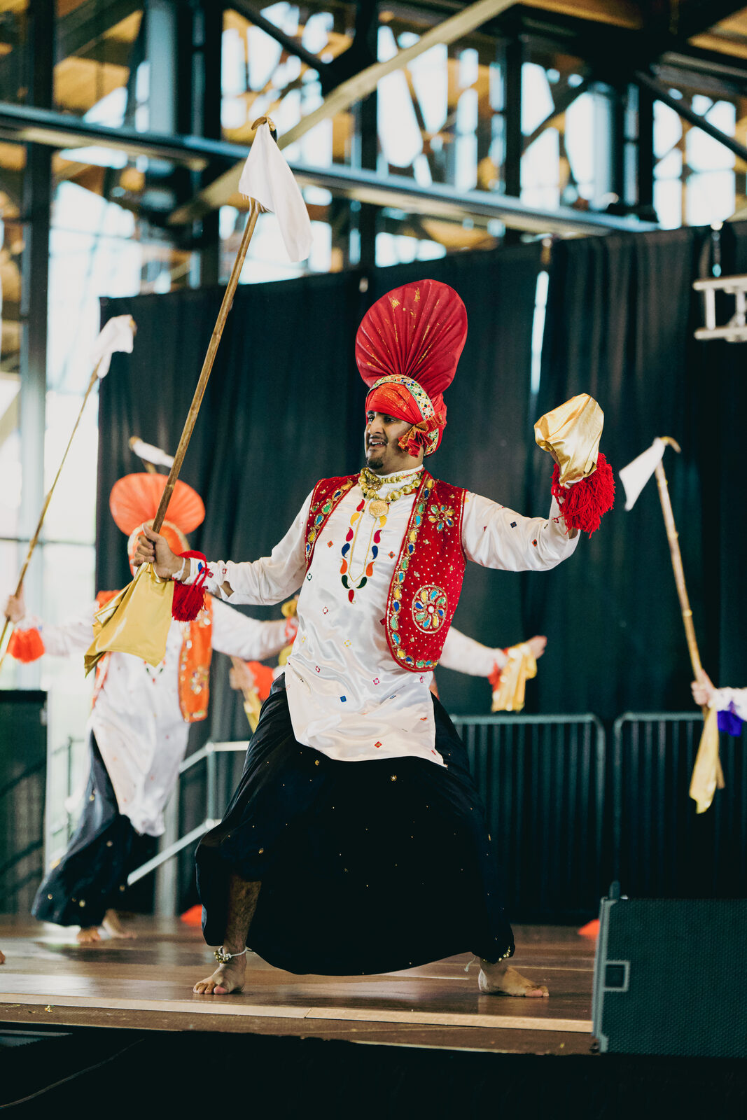 A Bhangra performer in an orange turban mid-kick with scissor props.