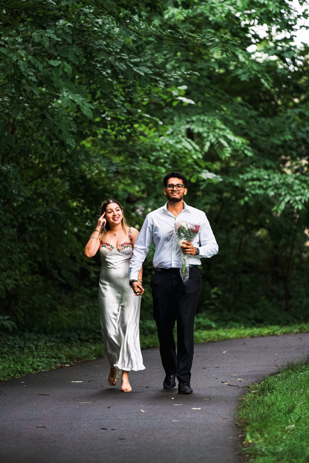 A couple walking down a wooded path, one holding a bouquet, moments before the surprise.