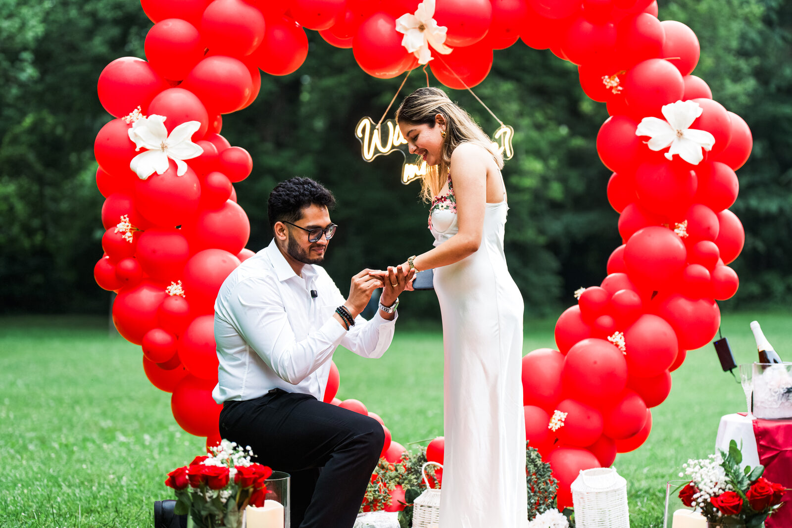 A man on one knee proposing under a red balloon heart arch reading Will You Marry Me.