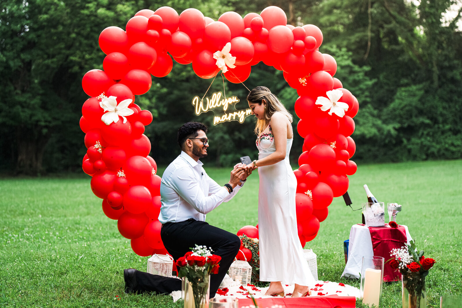 A close moment of the proposal under the balloon arch, placing the ring on her finger.